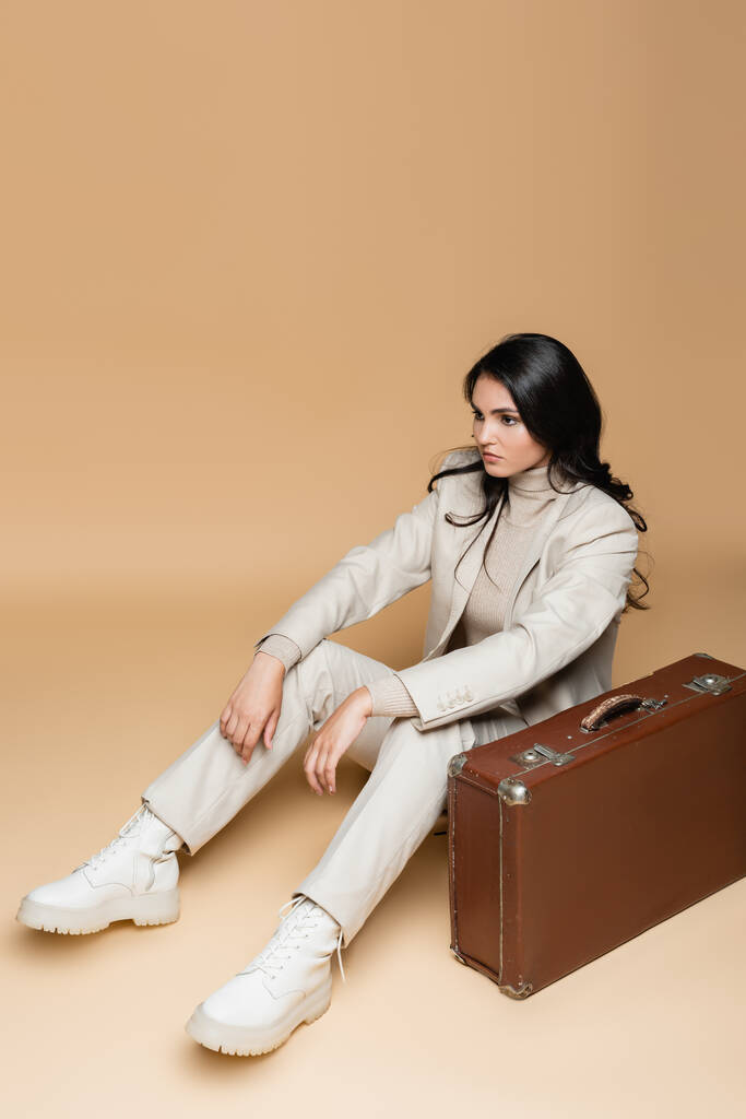 молодая женщина в костюме сидит возле винтажного чемодана на бежевом  - Фото, изображение