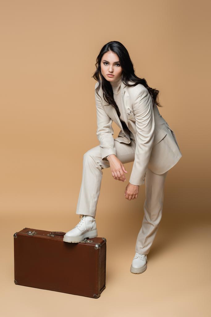 full length of young model in suit posing while standing near vintage suitcase on beige  - Photo, Image