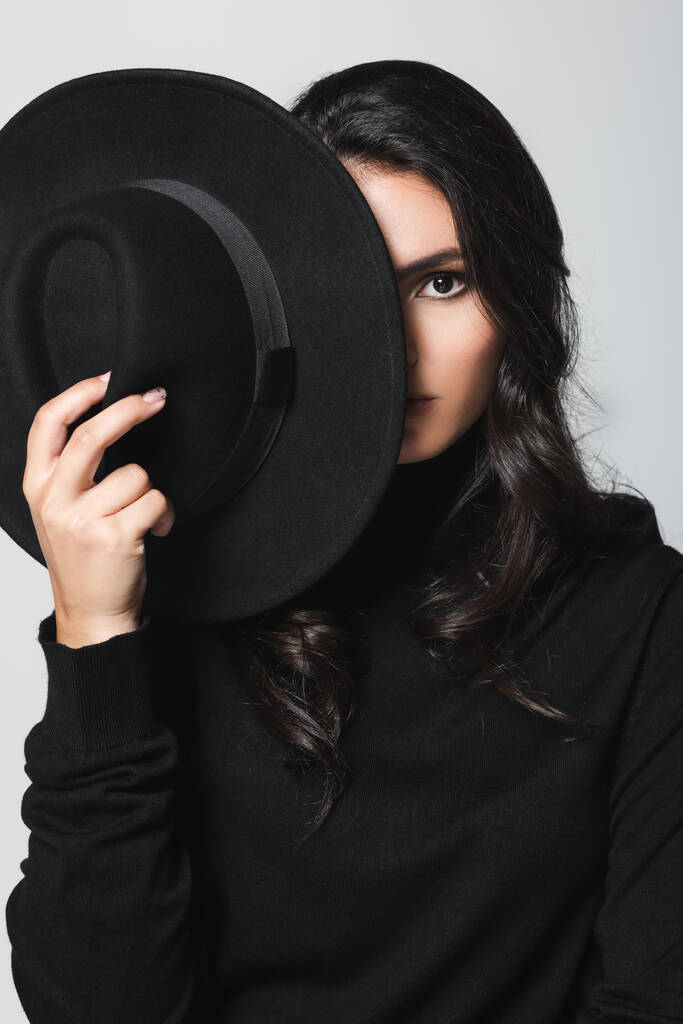 mladý model drží černý klobouk klobouk při zakrytí obličeje a při pohledu na fotoaparát izolované na šedé - Fotografie, Obrázek