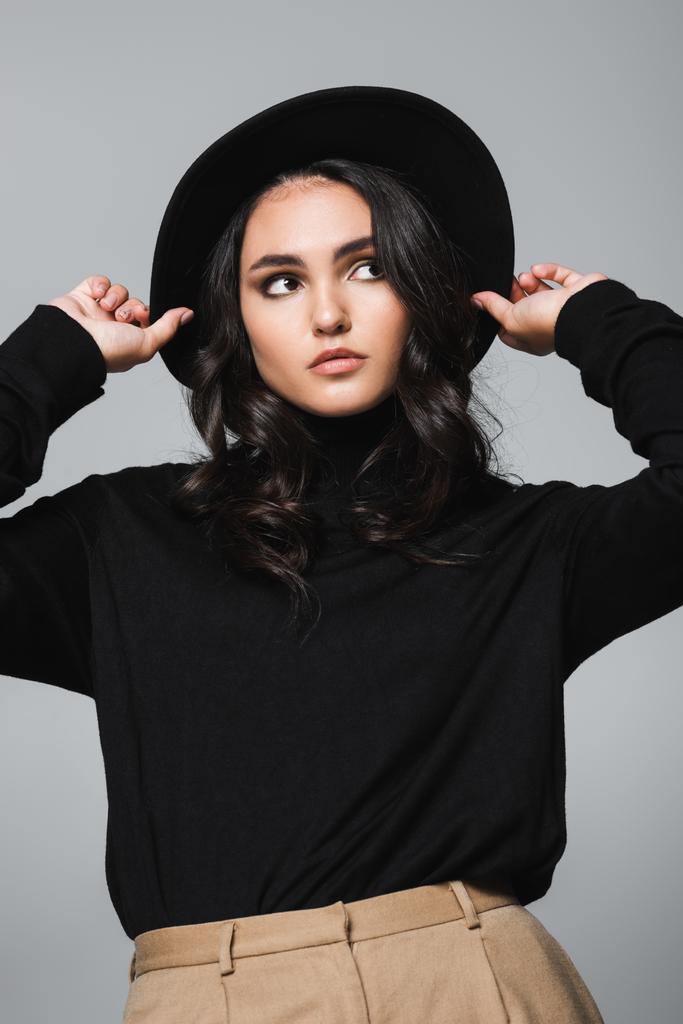 bruneta a mladý model úprava černé klobouk fedora a pohled pryč izolované na šedé - Fotografie, Obrázek