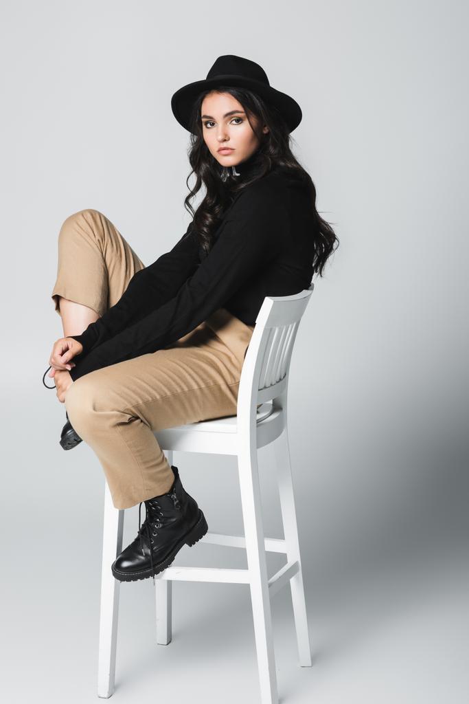 В полный рост молодая симпатичная женщина в федоровской шляпе смотрит на камеру, сидя на стуле на сером  - Фото, изображение
