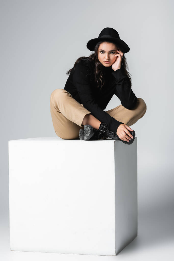 полная длина стильной модели в федоровской шляпе, смотрящей в камеру, сидя на белом кубе на сером  - Фото, изображение