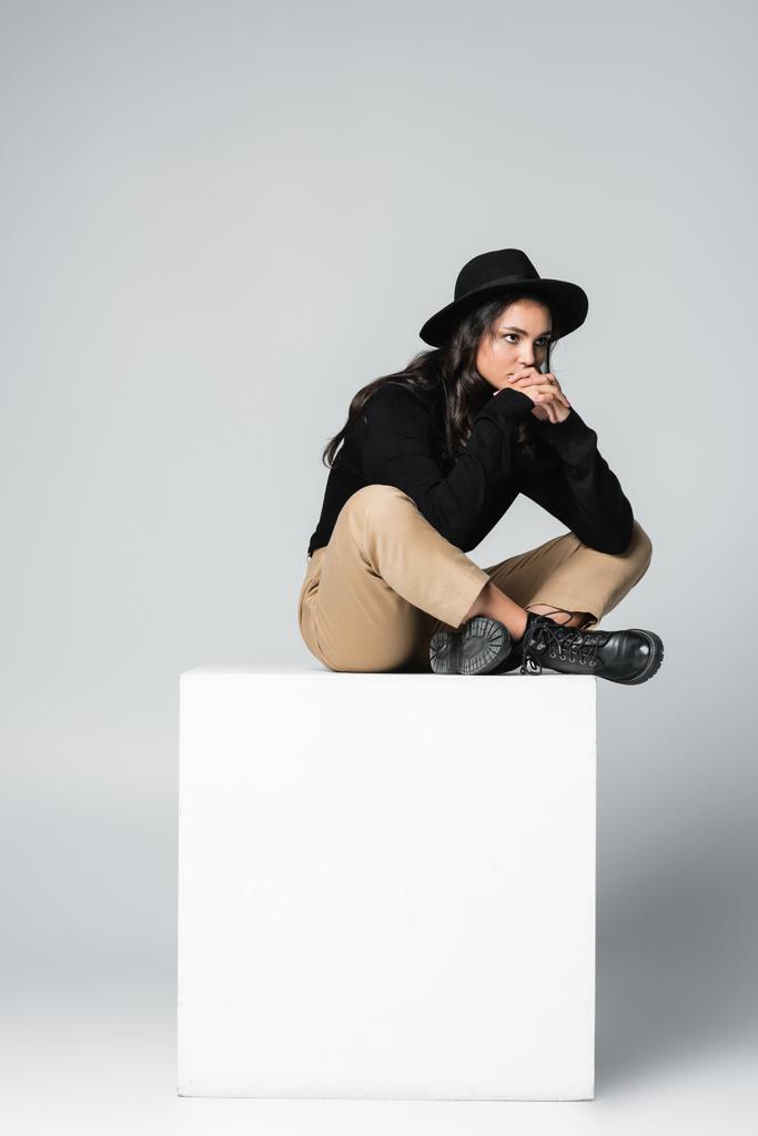 полная длина задумчивой модели в шляпе fedora сидя со скрещенными ногами на белом кубе на сером  - Фото, изображение