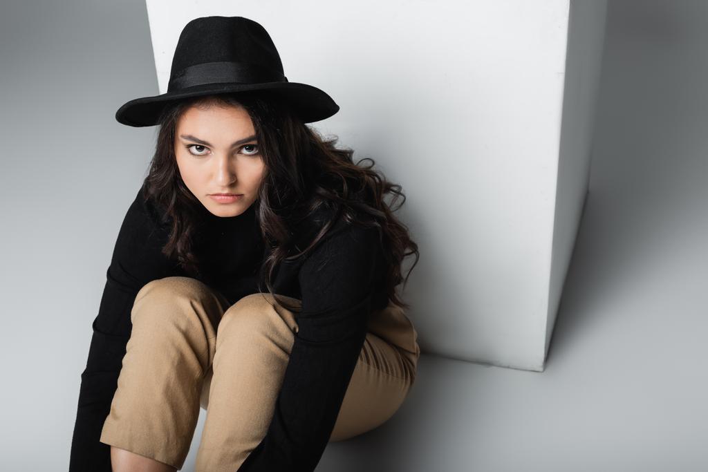 high angle view of stylish model in fedora hat looking at camera while sitting near white cube on grey  - Photo, Image