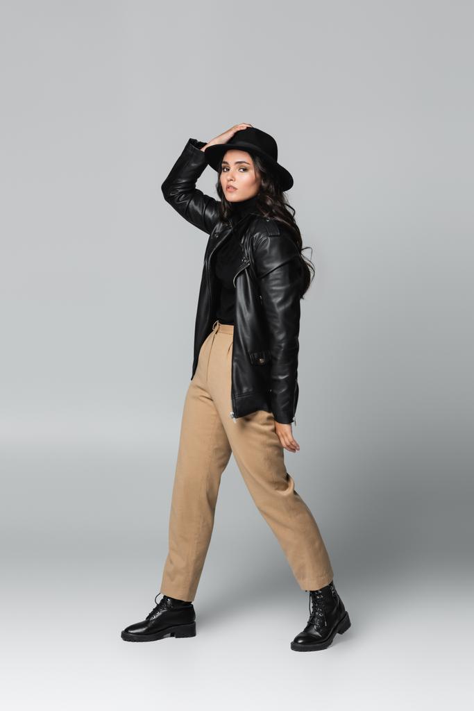 Trendmodel in voller Länge mit Fedora-Hut und schwarzer Lederjacke posiert auf grau  - Foto, Bild