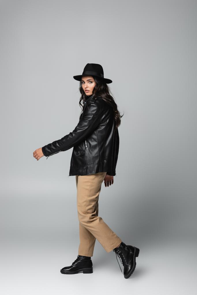 full length of trendy γυναίκα σε καπέλο fedora και μαύρο δερμάτινο μπουφάν ποζάρουν σε γκρι  - Φωτογραφία, εικόνα