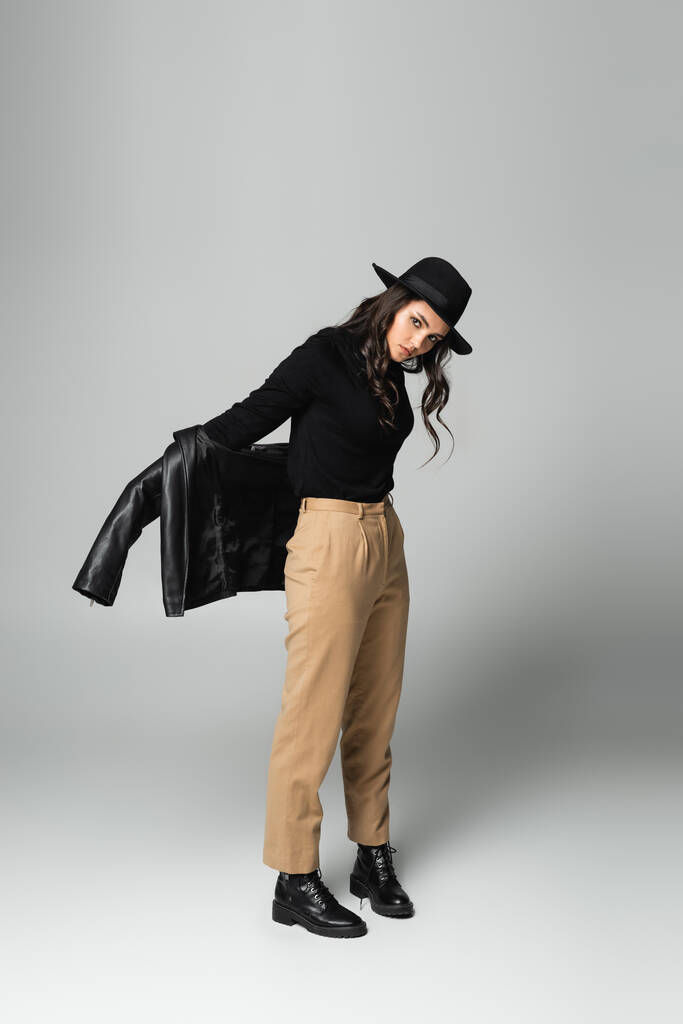 full length of stylish woman in fedora hat holding black leather jacket while posing on grey  - Photo, Image