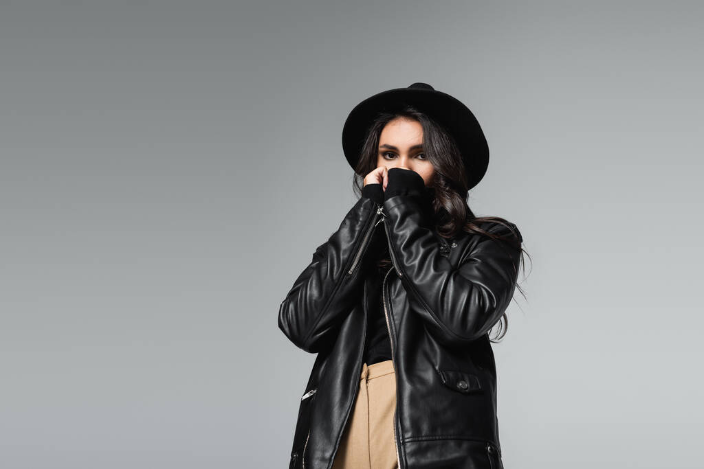 young woman in fedora hat and trendy leather jacket covering face with hands isolated on grey - Photo, Image
