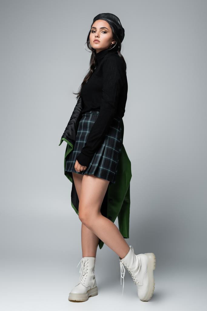 full length of young woman in green leather jacket, checkered skirt and black beret posing on grey - Photo, Image