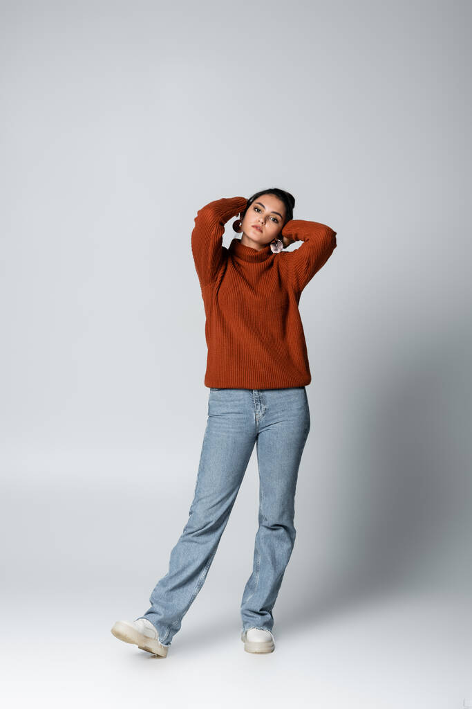 comprimento total da jovem mulher em camisola e jeans jeans posando em cinza - Foto, Imagem
