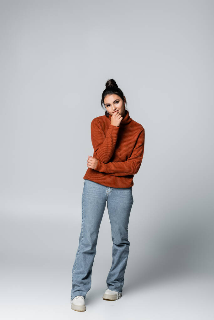 full length of smiling young woman in sweater and denim jeans posing on grey - Photo, Image