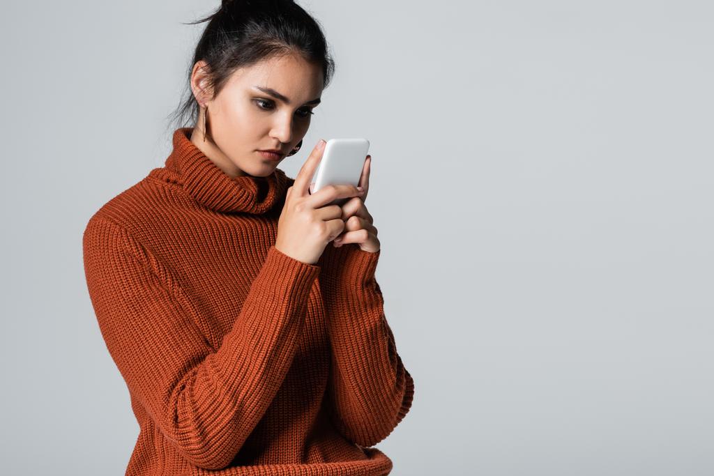 mujer joven en suéter de punto de mensajería en el teléfono inteligente aislado en gris - Foto, Imagen