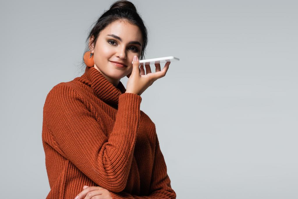 счастливая молодая женщина в вязаном свитере записывает голосовое сообщение на мобильный телефон, изолированный на сером - Фото, изображение