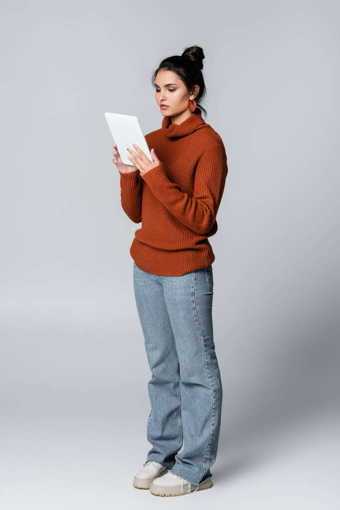 volledige lengte van de jonge vrouw in gebreide trui en jeans met digitale tablet op grijs  - Foto, afbeelding