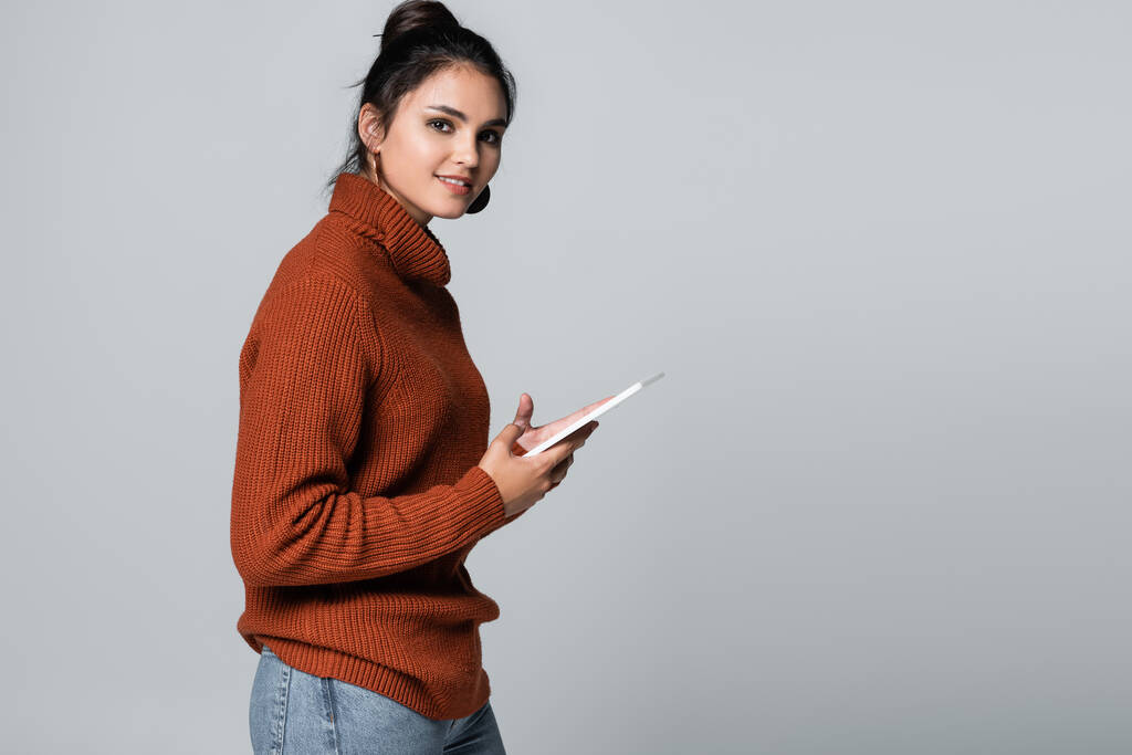 Gri üzerine izole edilmiş dijital tablet kullanan örgülü süveter giyen neşeli genç kadın.  - Fotoğraf, Görsel