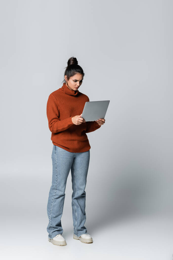 full length of young freelancer in knitted sweater and jeans using laptop on grey  - Photo, Image