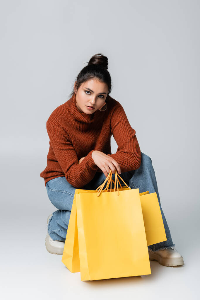 comprimento total da jovem mulher em suéter e jeans posando perto de sacos de compras amarelos em cinza  - Foto, Imagem
