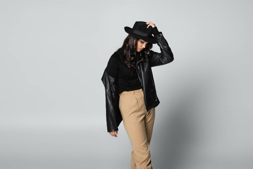 young model in black leather jacket and turtleneck adjusting fedora hat on grey - Photo, Image