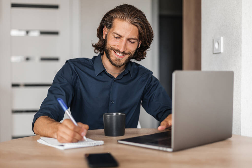 Homem satisfeito com um sorriso trabalha em um laptop e escreve em um caderno - Foto, Imagem