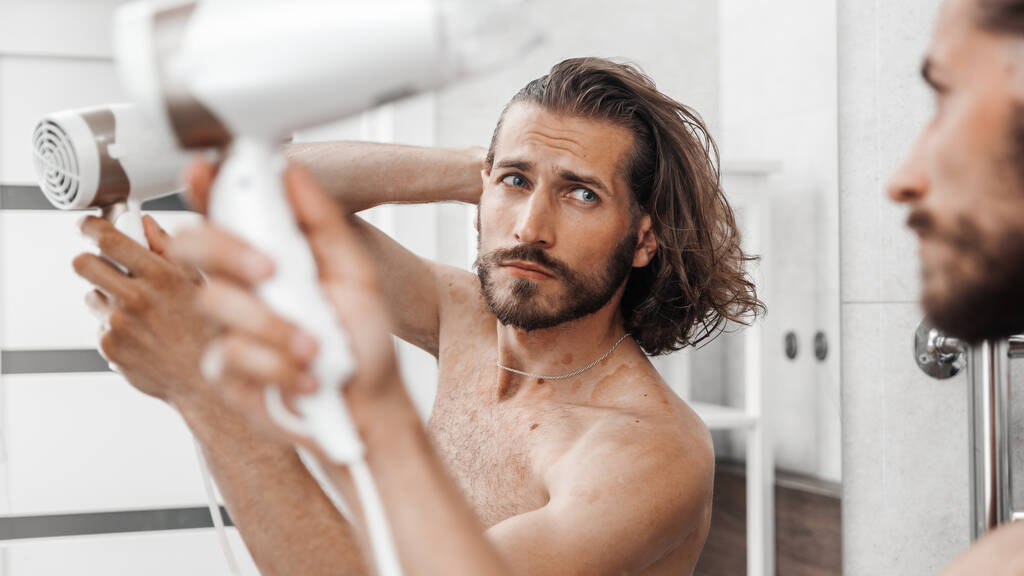 Hezký mladý muž se dívá do zrcadla a doma si suší vlasy fénem - Fotografie, Obrázek