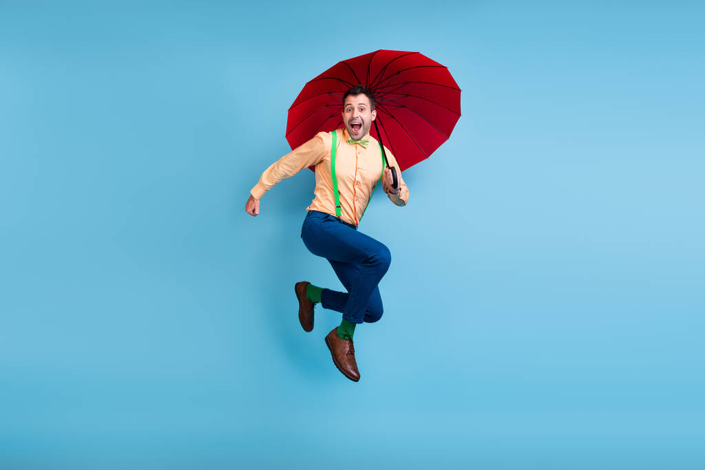 Full length photo of funny brunet man jump with parasol wear shirt trousers socks shoes isolated on blue color background - Photo, Image