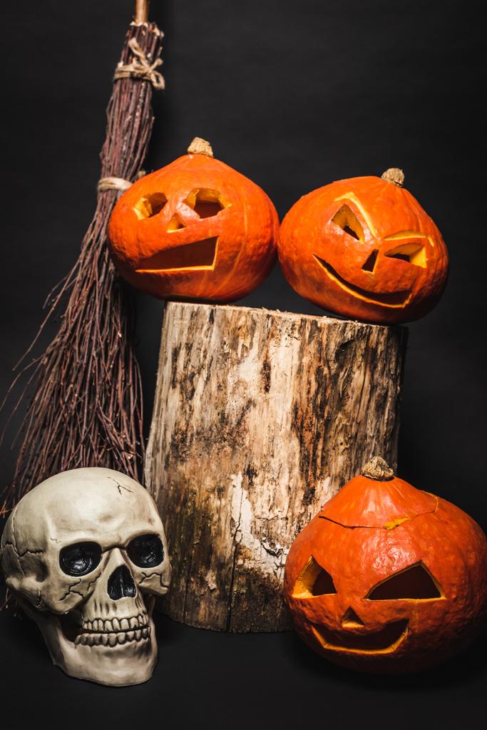 bezem bij oranje gesneden pompoenen op houten stomp en schedel op zwart - Foto, afbeelding