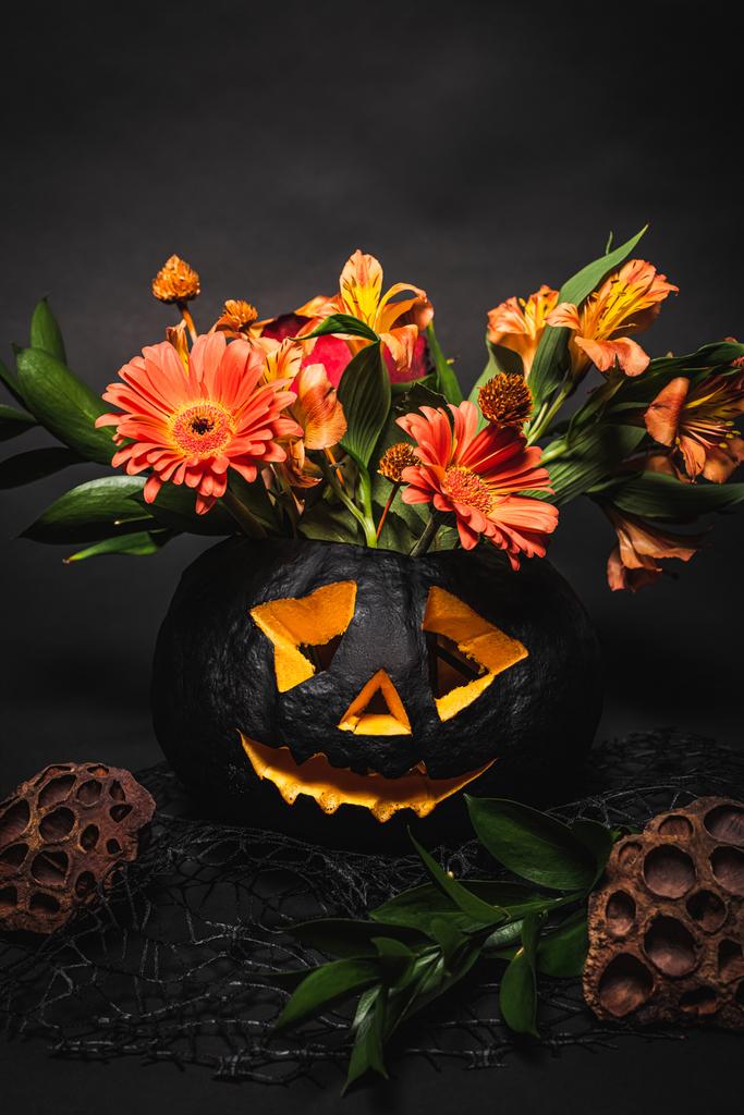 orange flowers with green leaves in spooky and carved pumpkin on black background  - Photo, Image