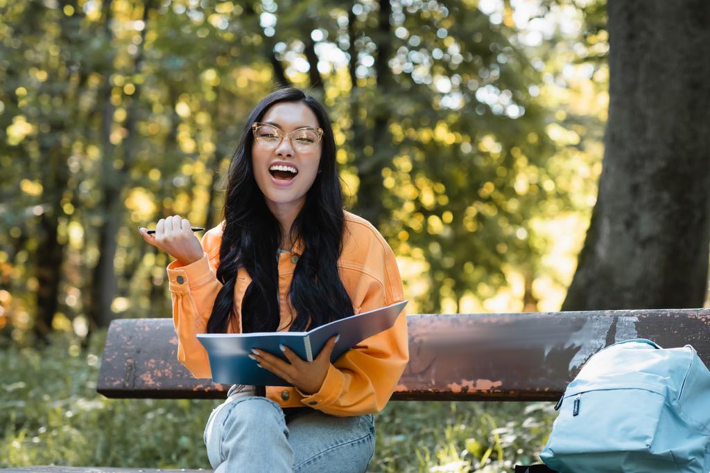emocionado asiático estudiante riendo en cámara mientras sentado en banco con notebook y pluma - Foto, Imagen