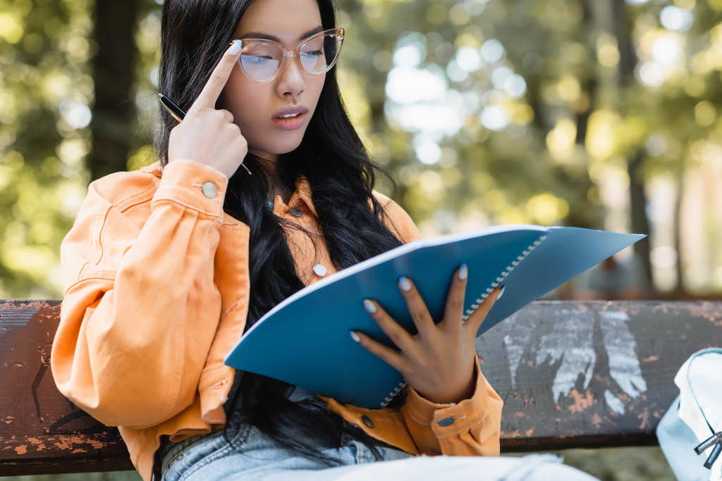 riflessivo asiatico studente regolazione occhiali mentre guardando in notebook in parco - Foto, immagini