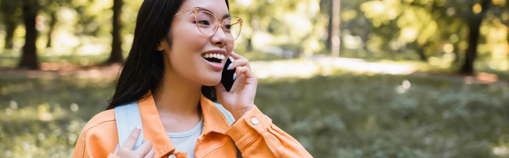 excitada mujer asiática en gafas hablando en el teléfono inteligente en el parque, pancarta - Foto, imagen