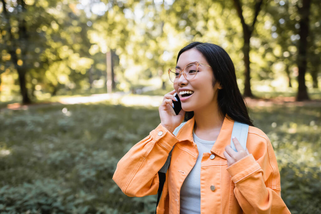 szczęśliwy Azji student w okulary i pomarańczowy kurtka rozmowy na telefon komórkowy w park - Zdjęcie, obraz