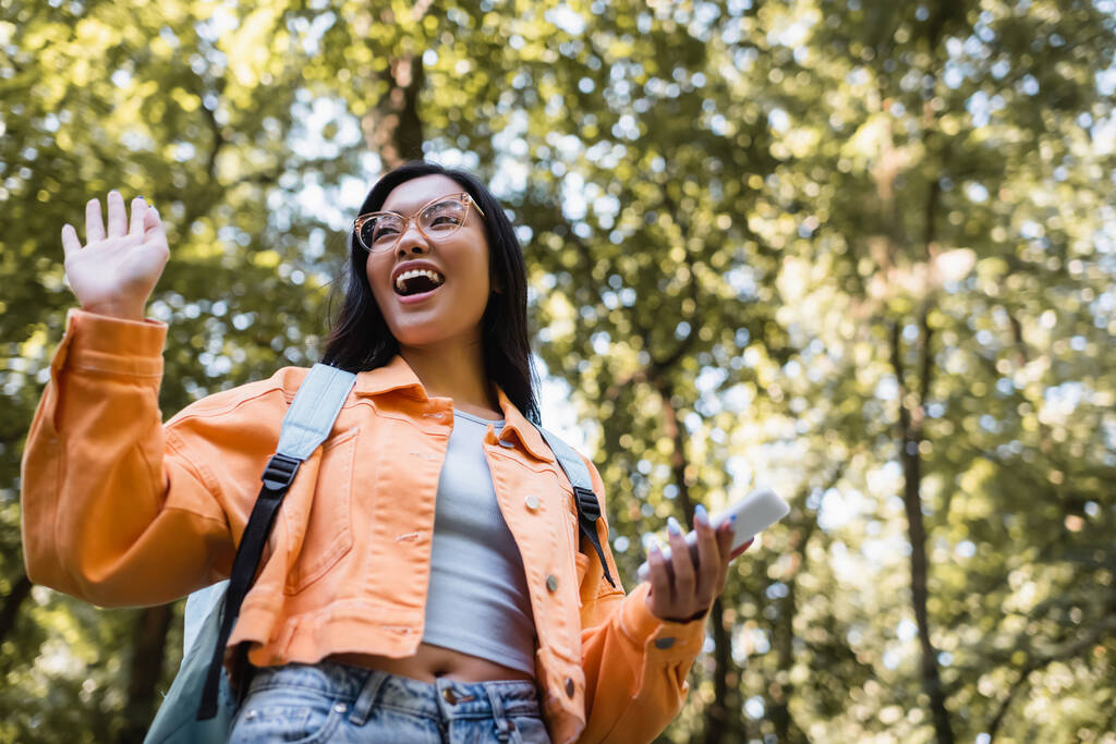 公園で手を振っているバックパックとスマートフォンを持つ興奮したアジアの学生の低角度ビュー - 写真・画像