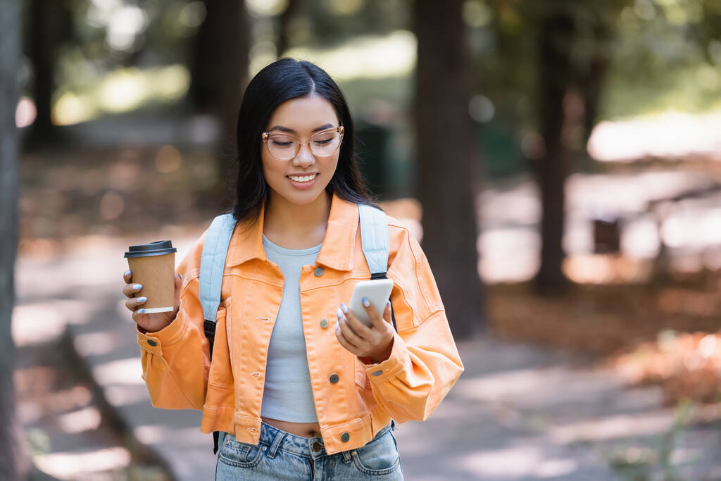 feliz asiático mulher olhando para celular enquanto segurando takeaway bebida ao ar livre - Foto, Imagem
