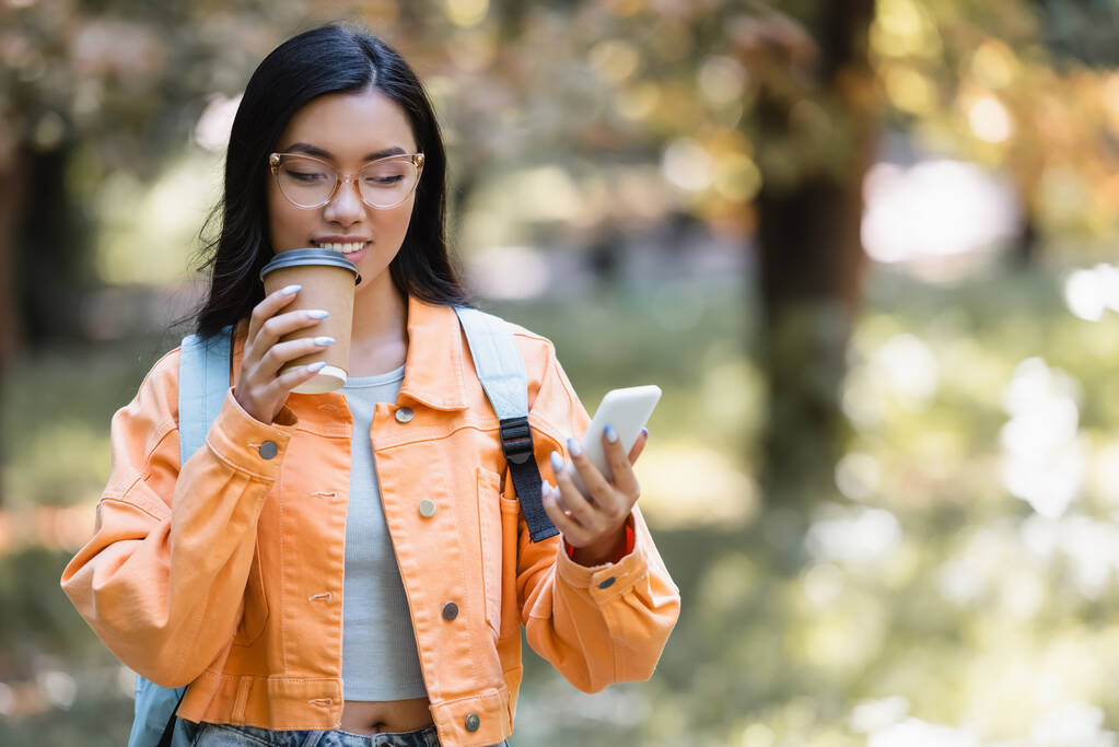 positieve aziatische student het drinken van koffie te gaan terwijl berichtgeving op mobiele telefoon in park - Foto, afbeelding