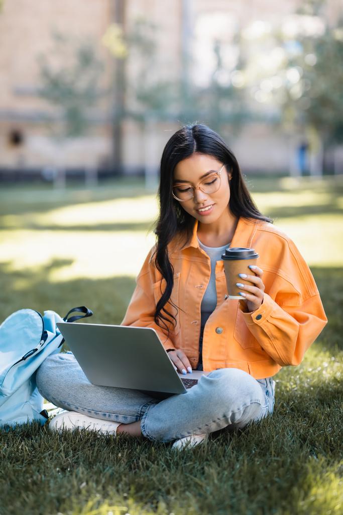 usmívající se asijské žena pomocí notebooku, zatímco sedí na trávníku s kávou jít - Fotografie, Obrázek