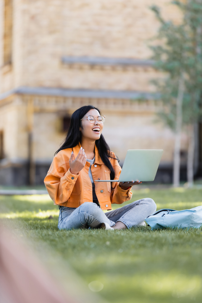 fröhliche asiatische Studentin lacht bei Videochat auf Rasen im Park mit geschlossenen Augen - Foto, Bild