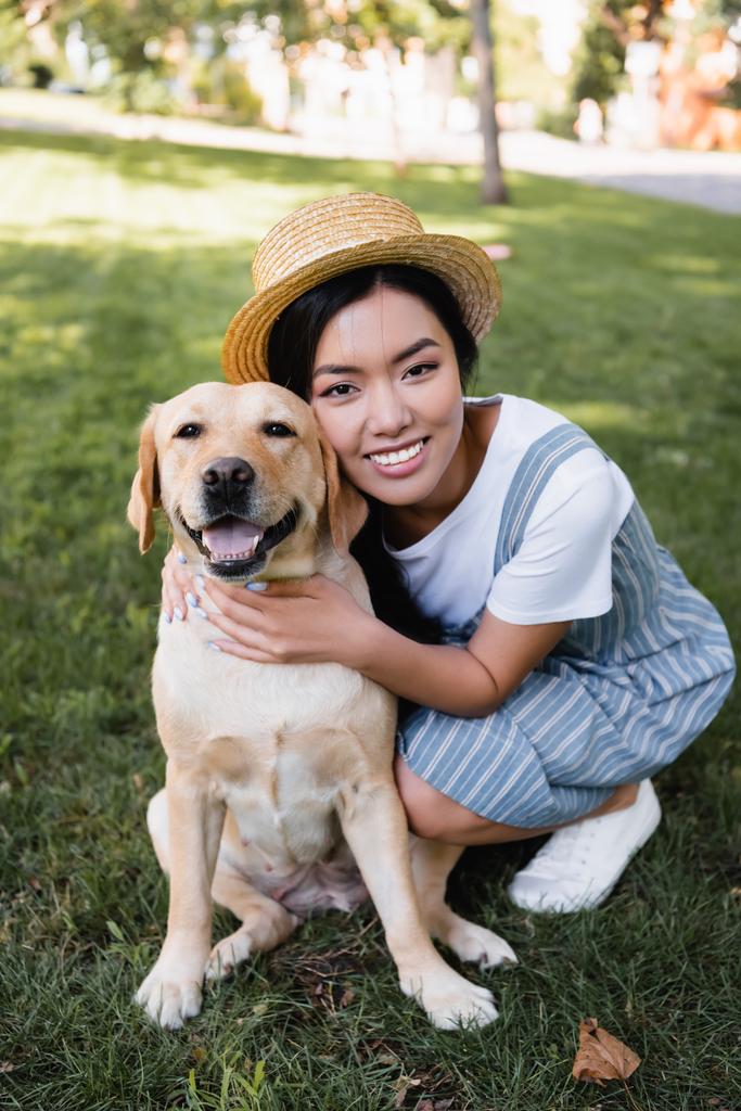młoda azjatycka kobieta uśmiecha się do kamery podczas przytulania psa w parku - Zdjęcie, obraz
