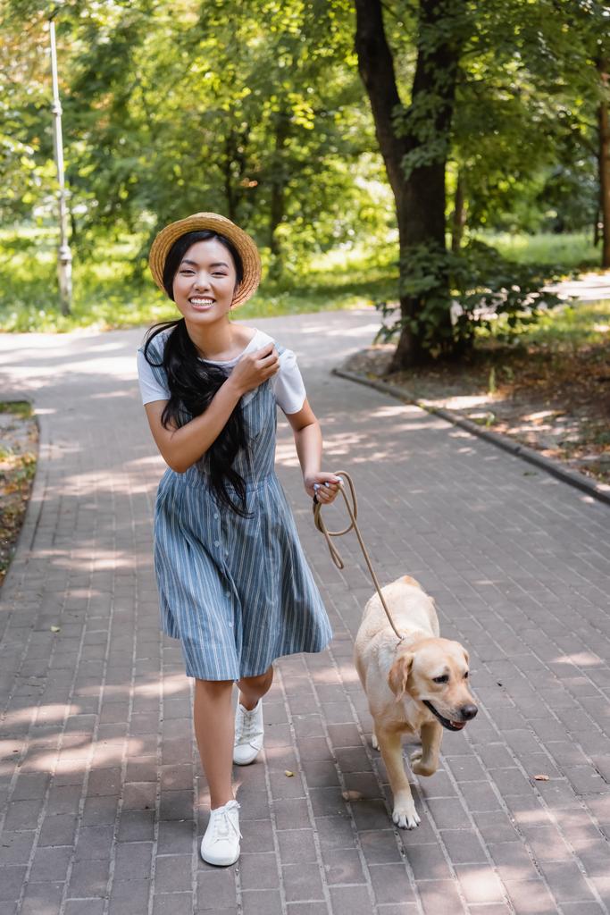 emocionado asiático mujer sonriendo en cámara mientras paseando con perro en parque - Foto, Imagen