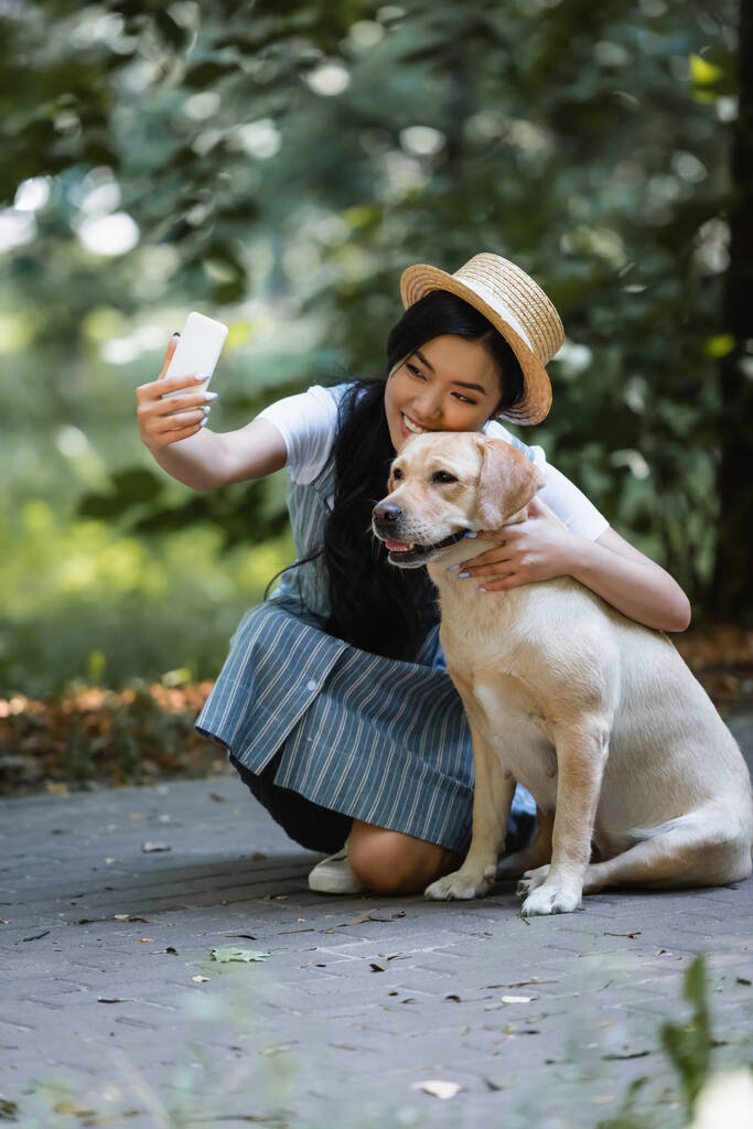cheerful asian woman taking picture on smartphone with labrador in park - Photo, Image