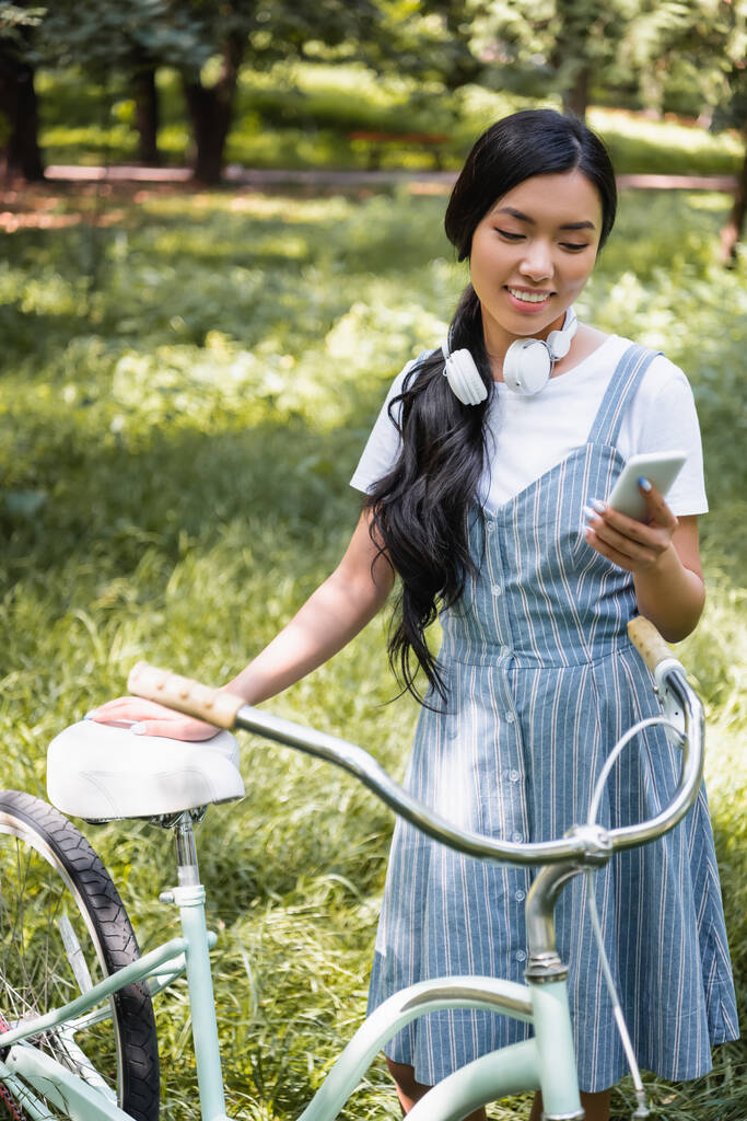 pozytywny Azji kobieta z słuchawki czat na telefon komórkowy w pobliżu rower w park - Zdjęcie, obraz