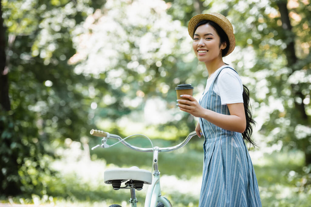 szczęśliwy Azji kobieta z na wynos drink patrząc w kamery w pobliżu rower w park - Zdjęcie, obraz