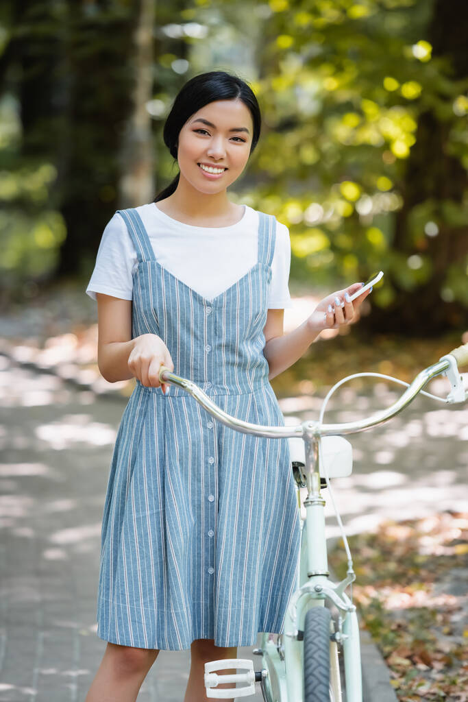 jovem mulher asiática com smartphone sorrindo para câmera perto de bicicleta no parque - Foto, Imagem