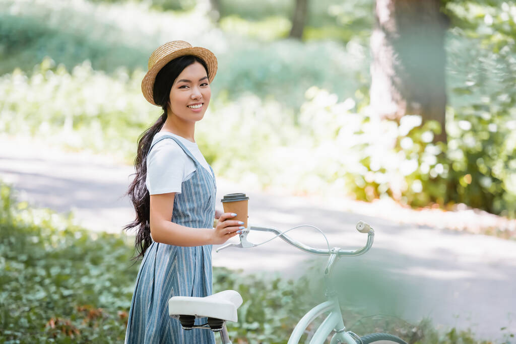 ストローハットの若いアジア系女性が公園の自転車の近くに行くためにコーヒーを持っていて - 写真・画像