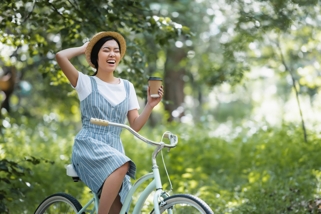 コーヒーを飲み公園で目を閉じたまま自転車で笑う陽気なアジアの女性 - 写真・画像