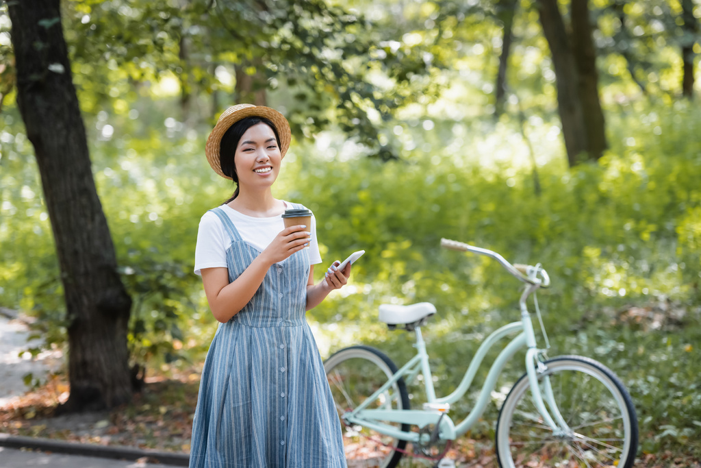 mujer asiática feliz con bebida para llevar y teléfono móvil mirando la cámara cerca de bicicleta en el parque - Foto, imagen