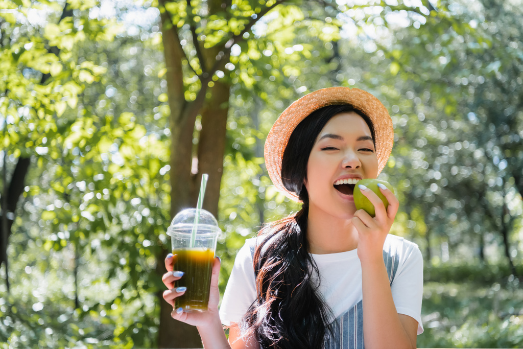 pleased asian woman with fresh smoothie biting juicy apple in park - Photo, Image