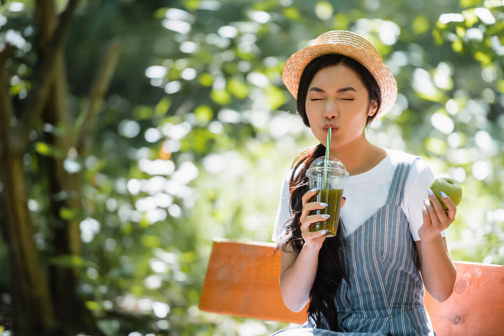 Gözleri kapalı Asyalı kadın parkta bankta taze smoothie içiyor. - Fotoğraf, Görsel