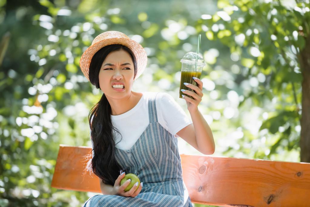 незадоволена азіатська жінка посміхається, дивлячись на смузі в пластиковій чашці
 - Фото, зображення