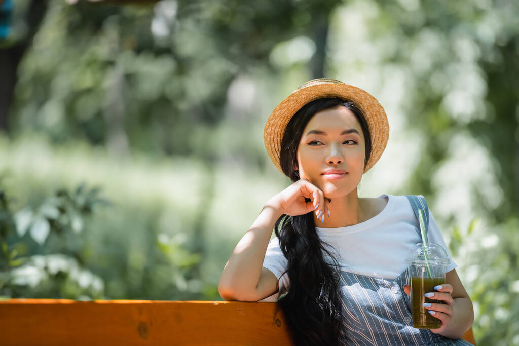 mujer asiática soñadora y sonriente en sombrero de paja sentado con batido fresco en el parque - Foto, imagen