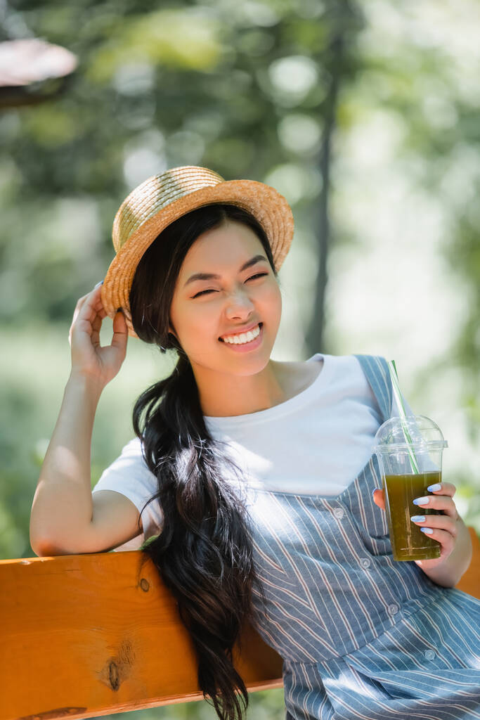 feliz asiático mujer con plástico taza de smoothie tocando paja sombrero en banco en parque - Foto, imagen